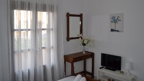 A hotel room with a bed, a television, a small table with a vase of flowers, a mirror, and a painting on the wall. - Floralia (Santorini)