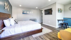 A bedroom with a wooden bed, white bedding, a gray air conditioner, and a small table with two stools. - White Caps Motel (Ventura)