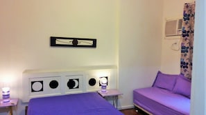 A room with a bed, a small table, a sofa, and a wall-mounted air conditioner.