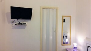 Ein Zimmer mit einem an der Wand befestigten Fernseher, einer Tür mit vertikalen Jalousien, einem Spiegel und einem Tisch mit einer Lampe.