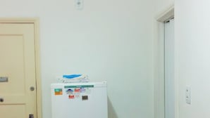 Ein kleiner Raum mit einem weißen Kühlschrank, einem violetten Sessel, einer weißen Tür und einer weißen Wand.