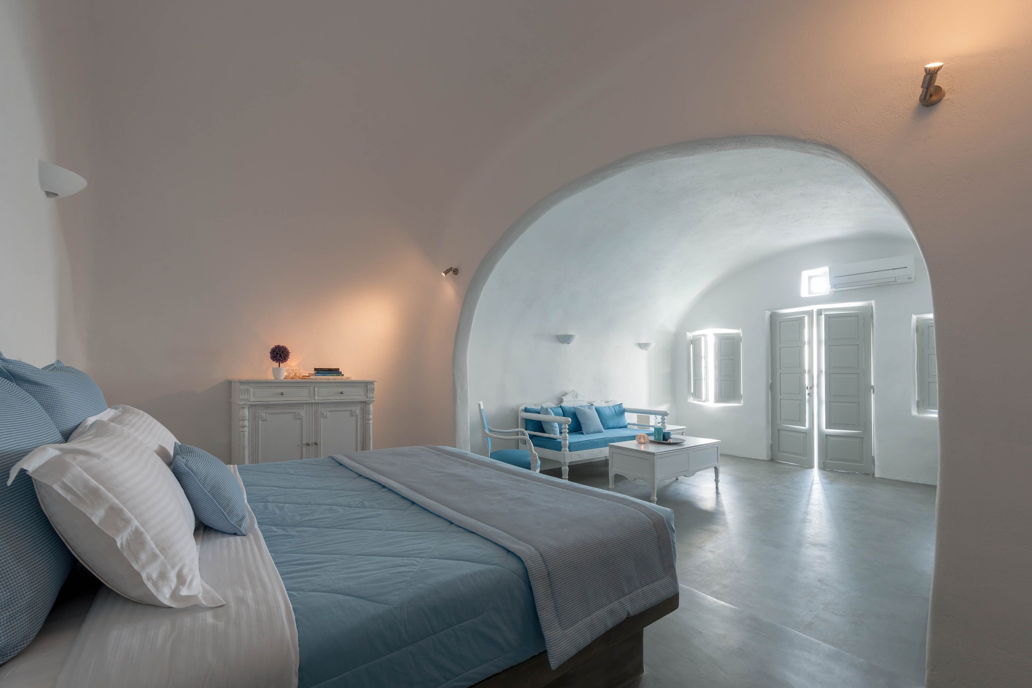 A bedroom with a bed, a nightstand, a chair, and a sofa. There is a large arched doorway leading to another room.