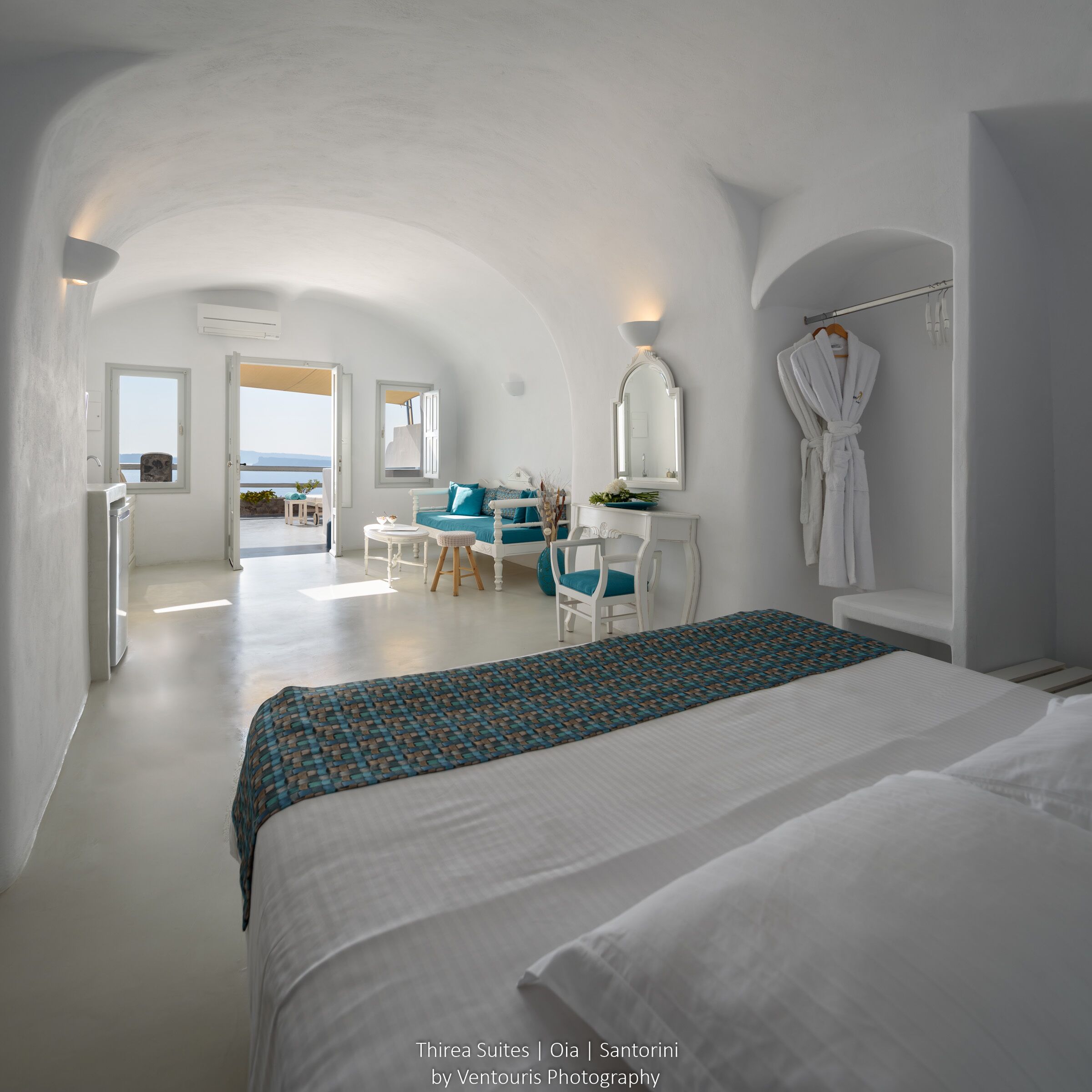 Une chambre à coucher moderne, aux couleurs claires et dominée par le blanc, avec un grand lit, un bureau et une chaise. Une porte ouverte offre une vue sur la mer.