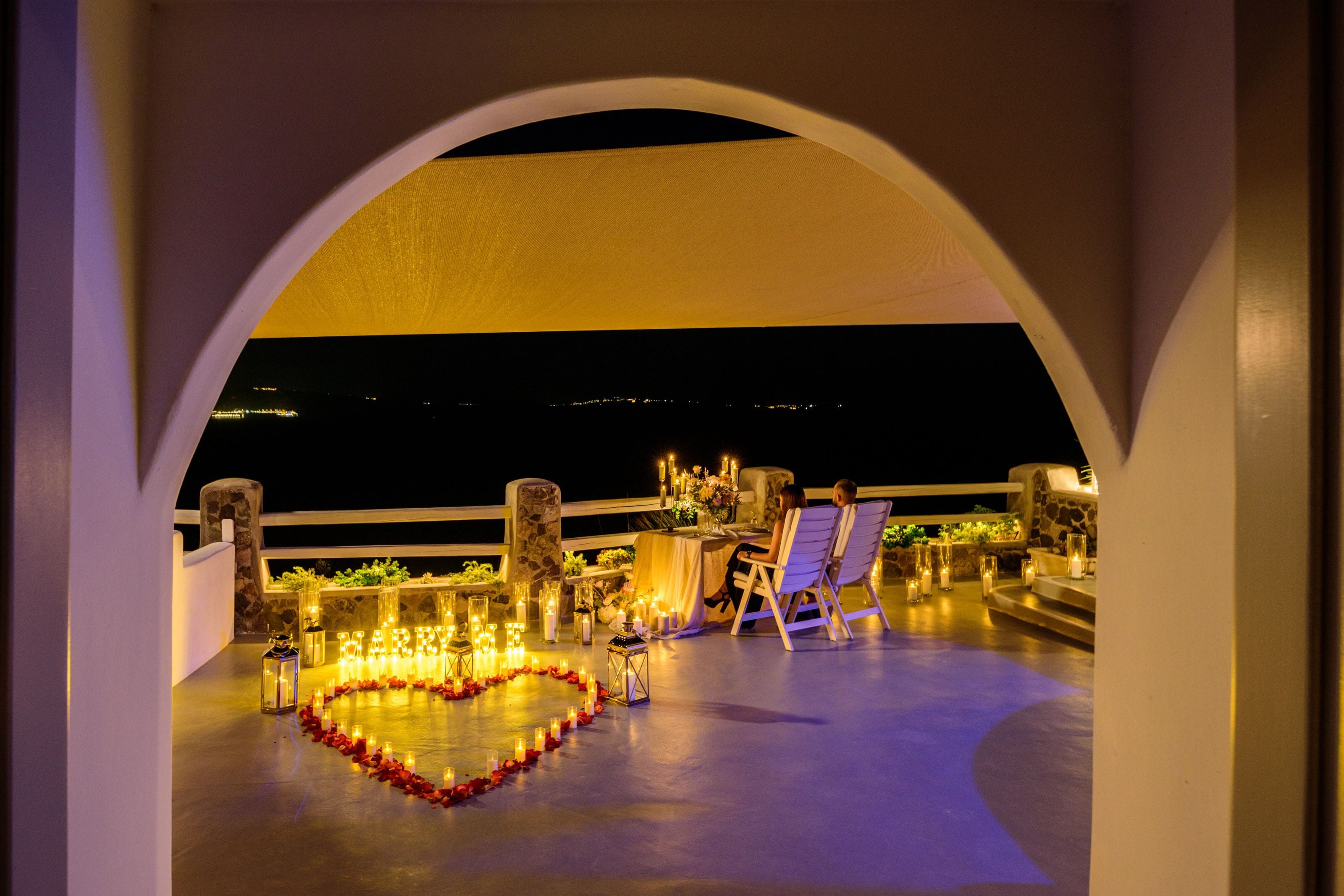 An outdoor seating area with a heart-shaped arrangement of candles, a table with chairs, and a view of the city at night.