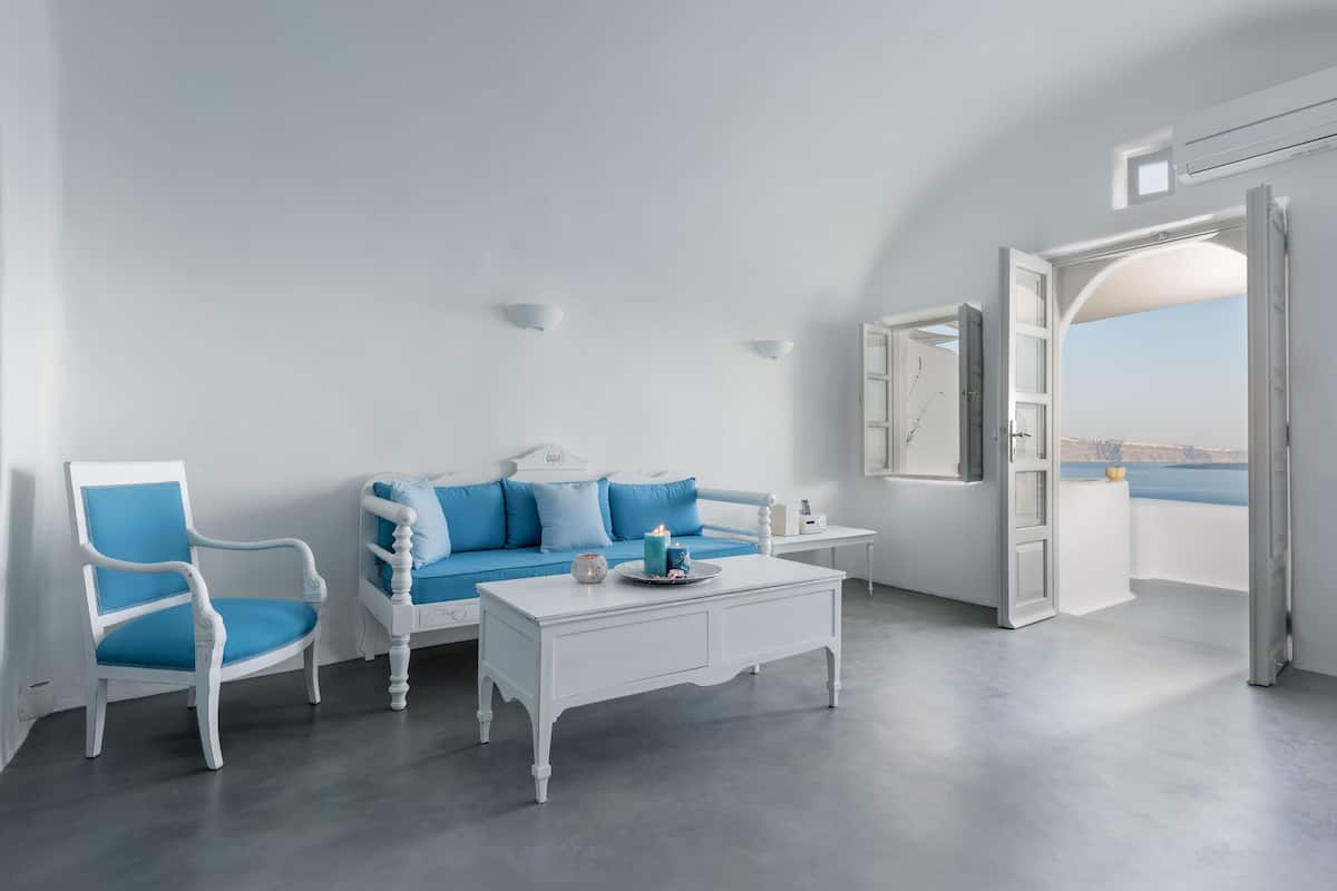 A room with a white sofa and chair, a small table, and a view of the sea through an open door.