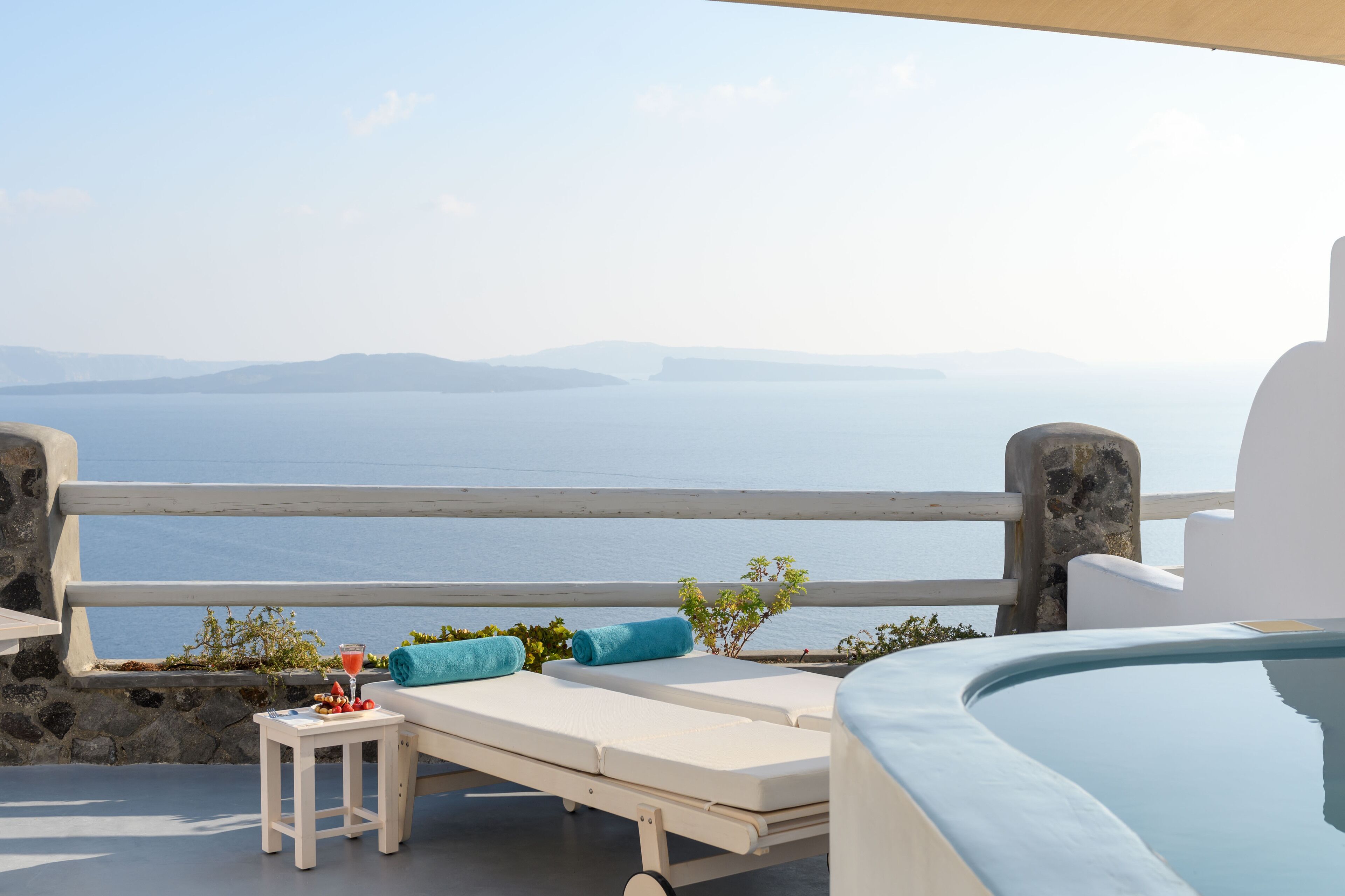 Un balcon ensoleillé avec un fauteuil blanc, une petite table sur laquelle sont posés une boisson et des fruits, et une vue sur la mer et les montagnes lointaines.