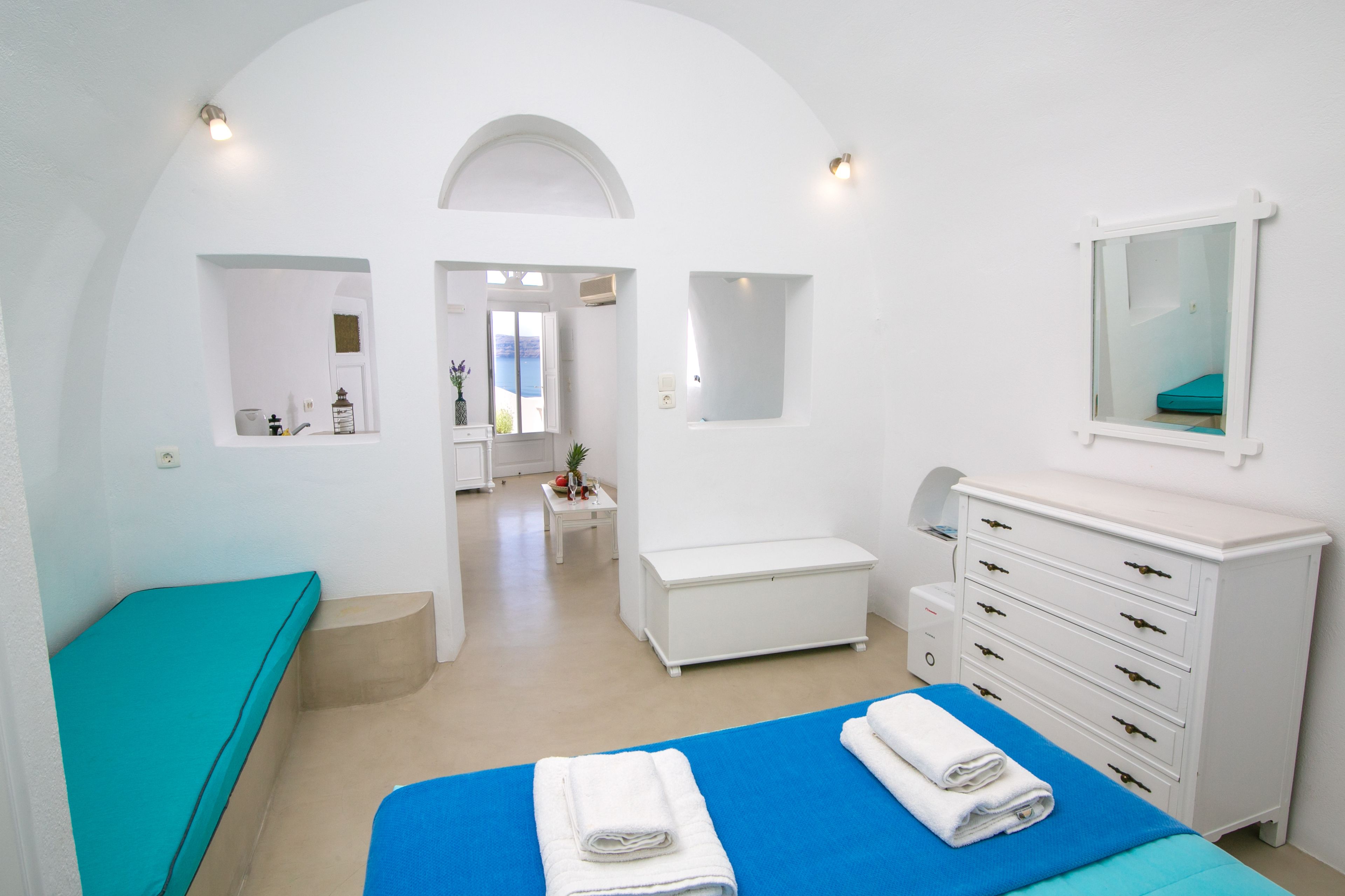 A modern, white-themed bedroom with a bed, a bench, a dresser, and a small table with a plant.