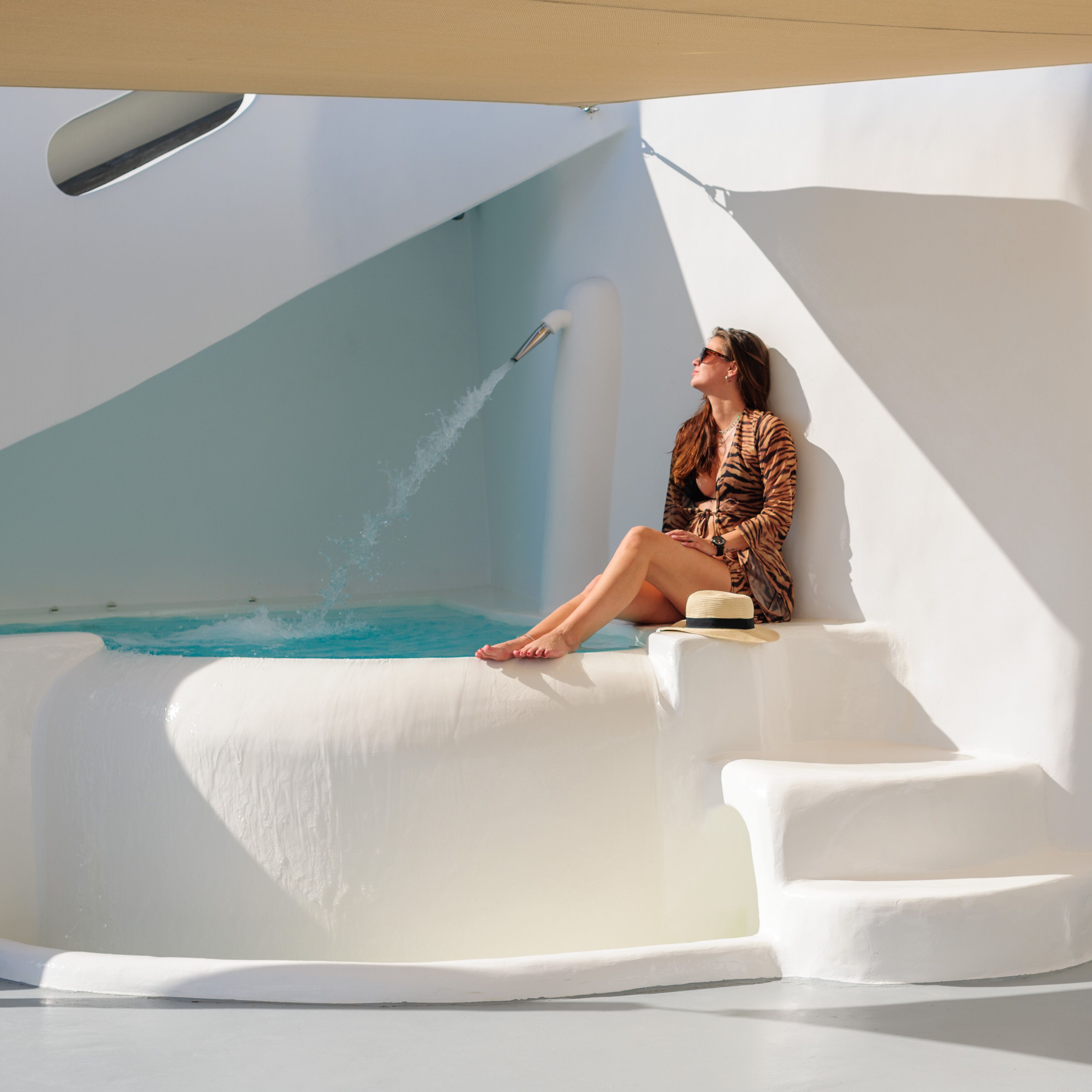 Une personne est assise au bord d’une piscine dont l’architecture moderne se distingue par un élément aquatique original et un parasol.