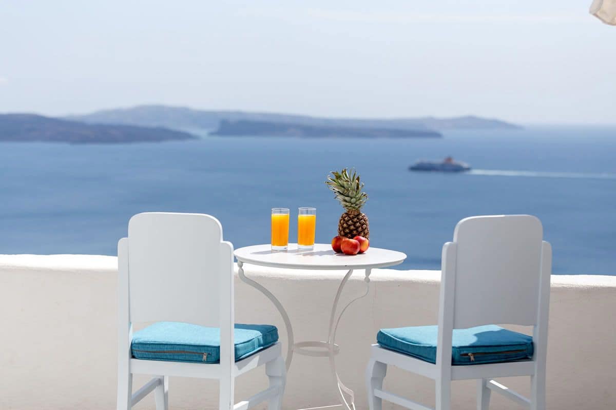 A balcony with two white chairs and a small table, featuring a view of the sea and distant islands.