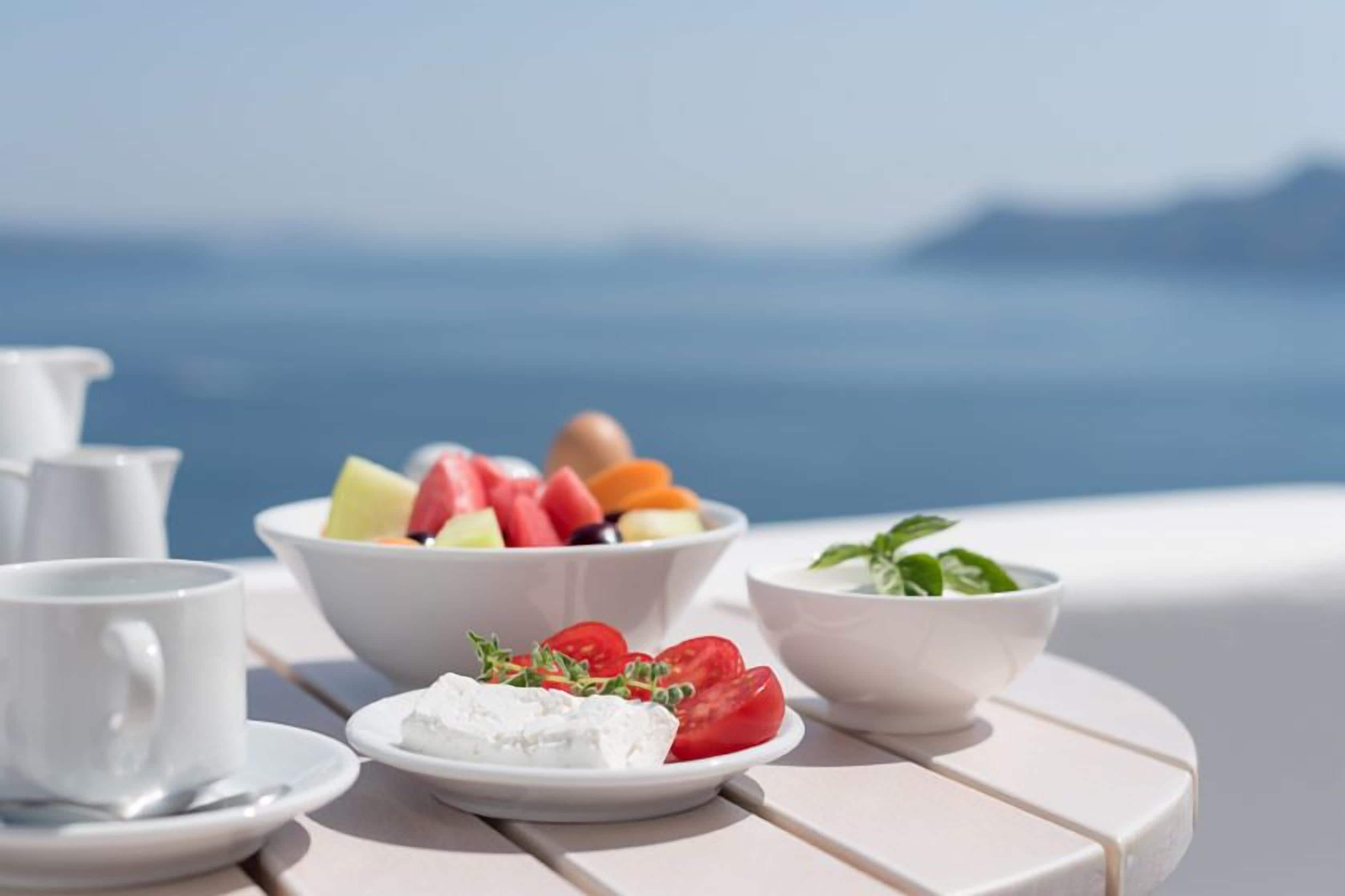 A table with a bowl of fruit salad, a plate of cheese, and a bowl of greens, set against a scenic view.