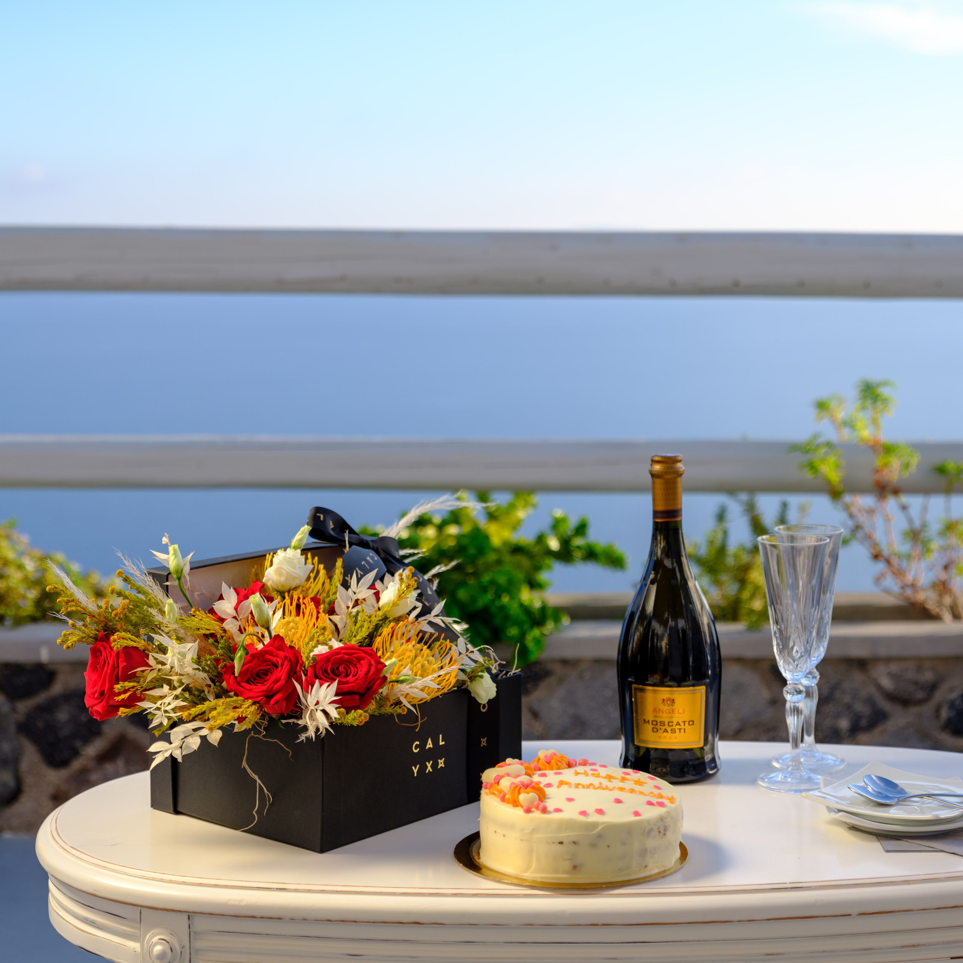 A table with a cake, a bottle of wine, and a bouquet of flowers on a balcony overlooking a body of water.