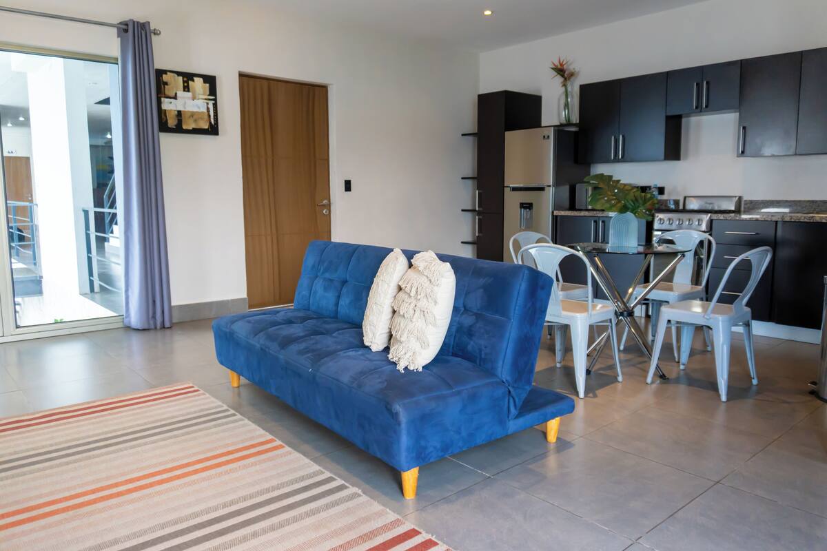 A modern living room with a blue sofa, a dining table with chairs, and a kitchen area with dark cabinets and a stove.