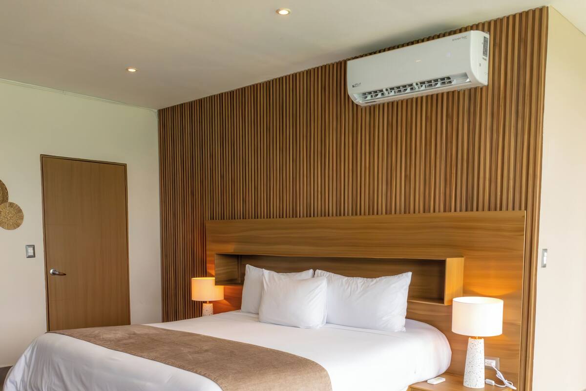 A hotel room with a large bed, wooden headboard, bedside lamps, and an air conditioning unit.