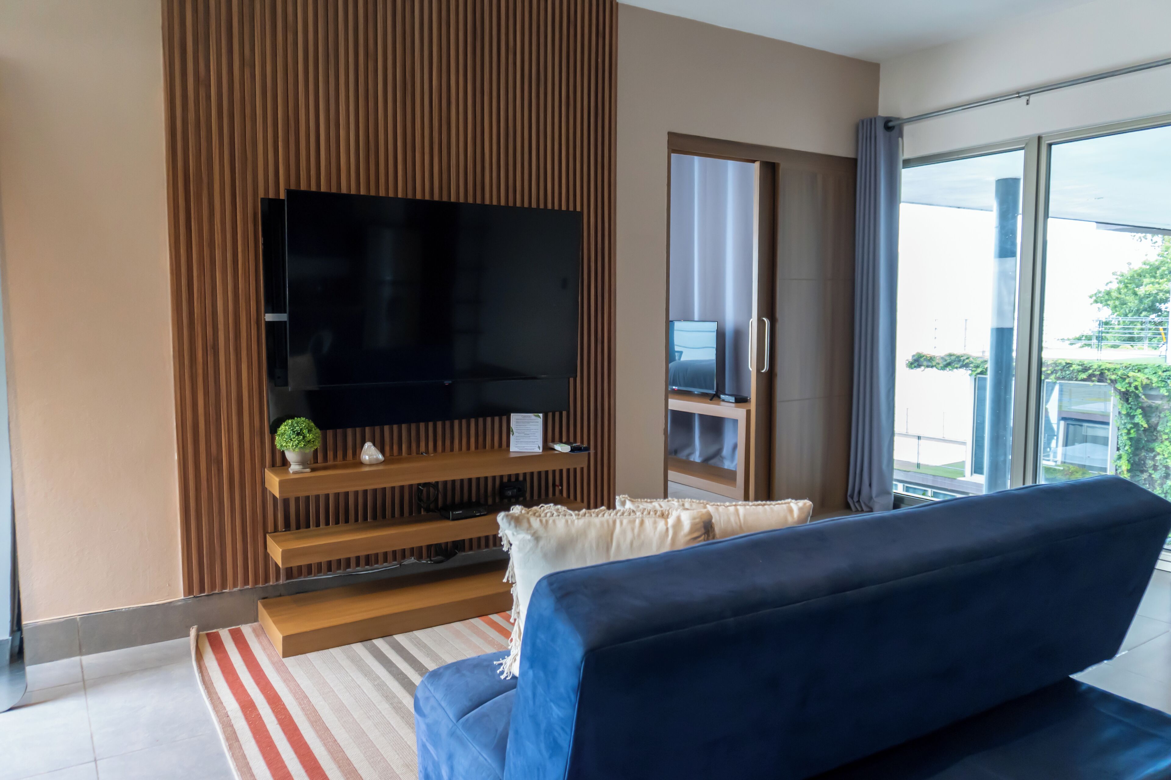 Un salon moderne doté d’un grand téléviseur à écran plat fixé sur un mur en panneaux de bois, d’un canapé bleu et d’une grande fenêtre offrant une vue sur l’extérieur.