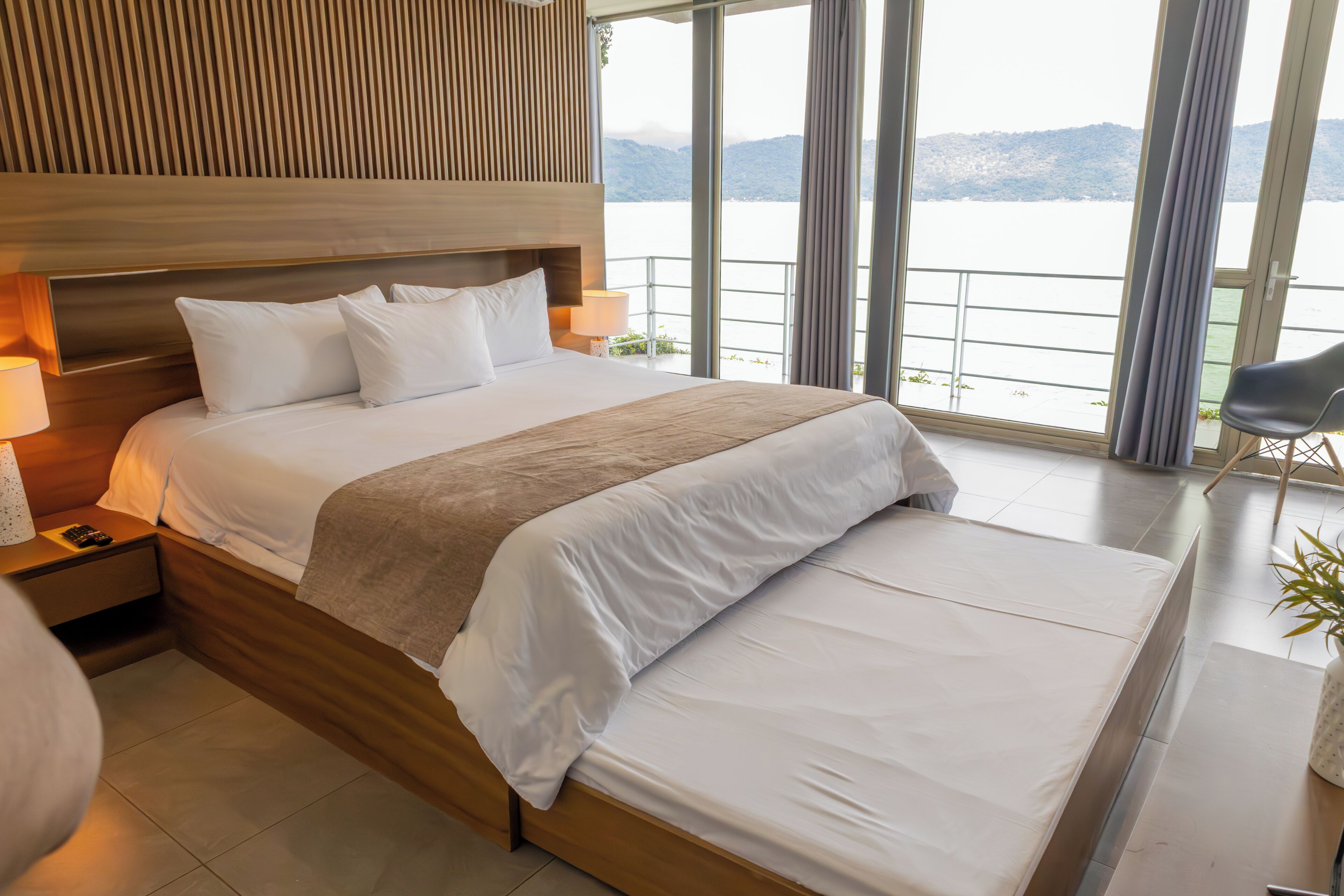 A modern hotel room with a large bed, wooden headboard, and a view of a lake through large windows.