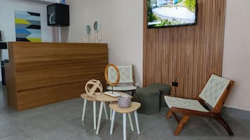 Un área de recepción con un mostrador de madera, un televisor montado en la pared y un espacio de descanso con sillas de mimbre y un banco acolchado.