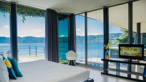 A bedroom with a large bed, a TV, and a balcony with a view of the water and mountains.