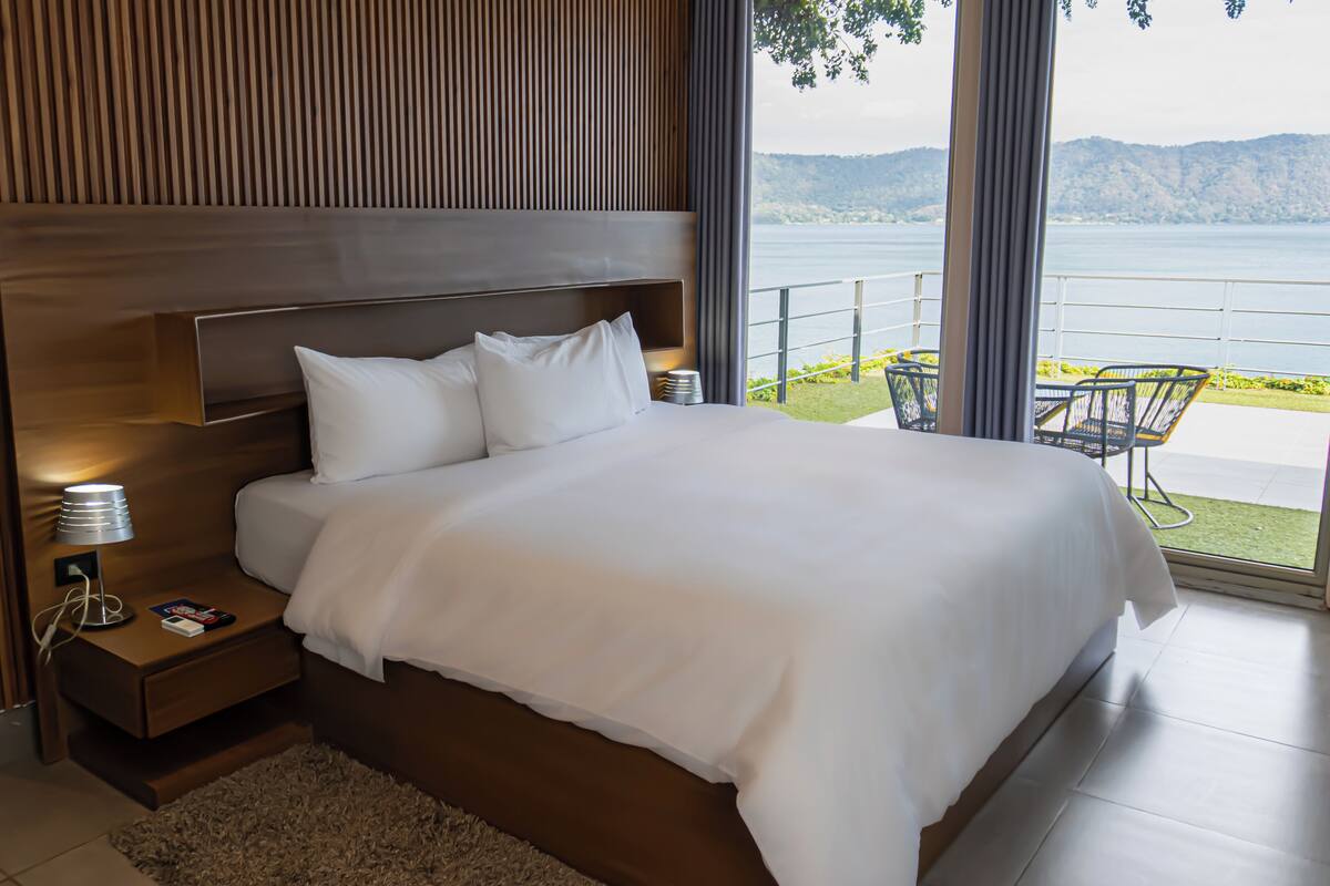 A modern bedroom with a large bed, wooden headboard, bedside table with lamp, and a view of a lake and mountains.