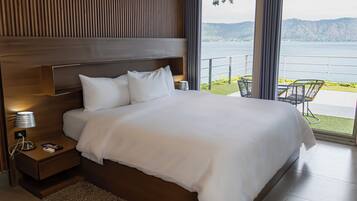 Un dormitorio moderno con una cama grande, cabecera de madera, mesita de noche con lámpara y vistas a un lago y montañas.