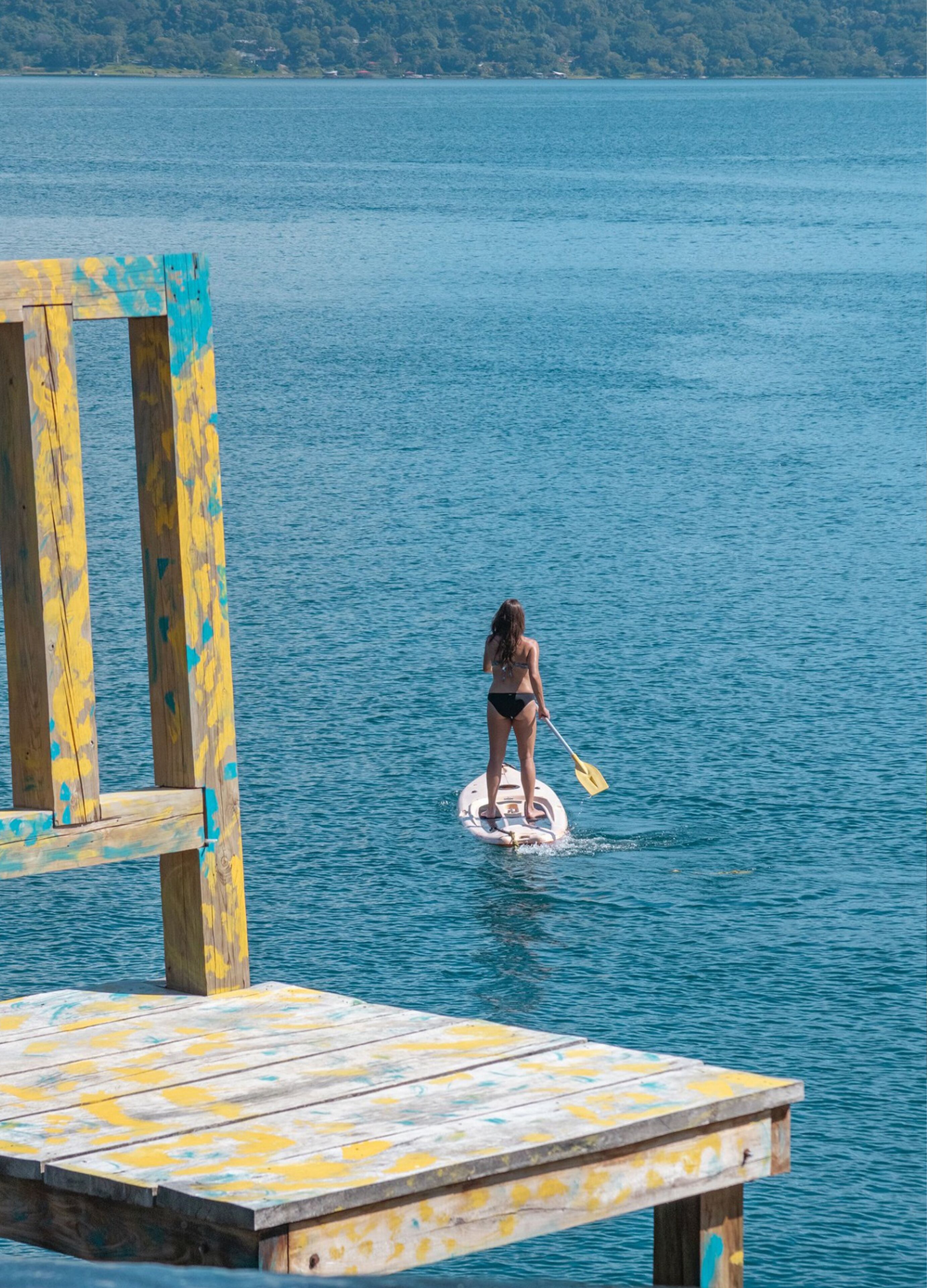 Une personne fait de la planche à pagaie sur une jetée en bois qui s’avance sur une étendue d’eau calme.