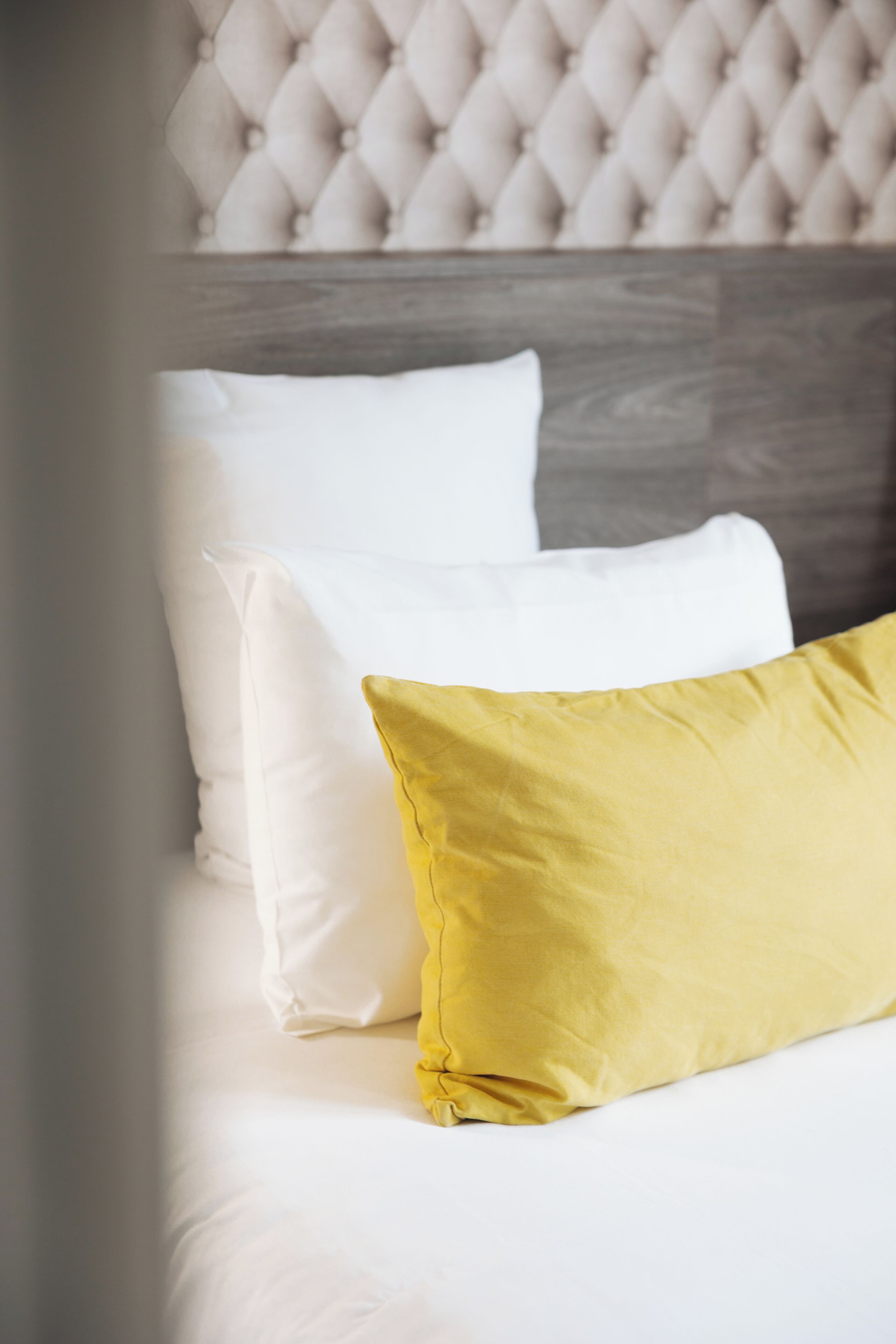 Una cama con almohadones blancos y amarillos, un cabecero tapizado y otro de madera.