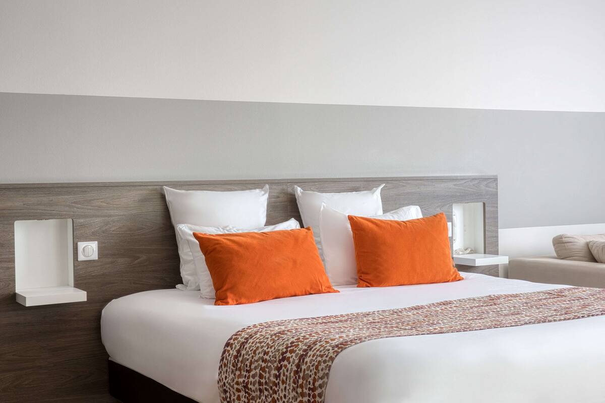 Ein modernes Hotelzimmer mit einem großen Bett, weißer Bettwäsche und zwei orangen Kissen. Ein Nachttisch mit Lampe und einer kleinen Ablagefläche ist ebenfalls vorhanden.