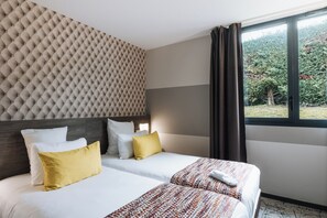 Habitación de hotel con cabecera capitonada, un ventanal con cortinas, cama con ropa de cama blanca y amarilla, y una manta estampada.