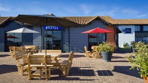 Een terras met houten tafels en banken, een grote parasol en potplanten.