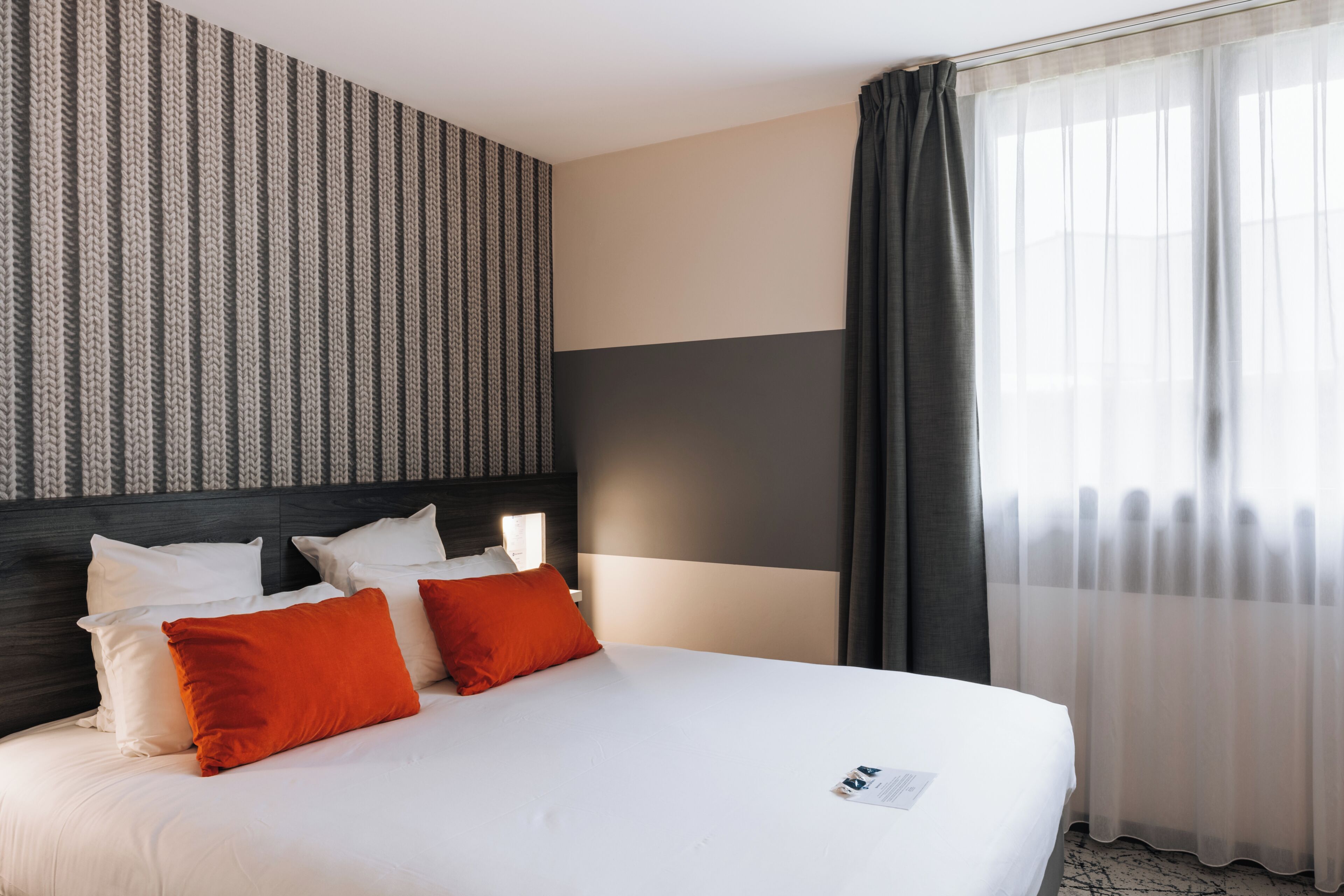 A hotel room with a bed, pillows, a headboard, a window with curtains, and a wall with a striped pattern.