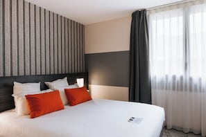 Una habitación de hotel con cama, almohadas, cabecera, ventana con cortinas y pared con diseño a rayas.