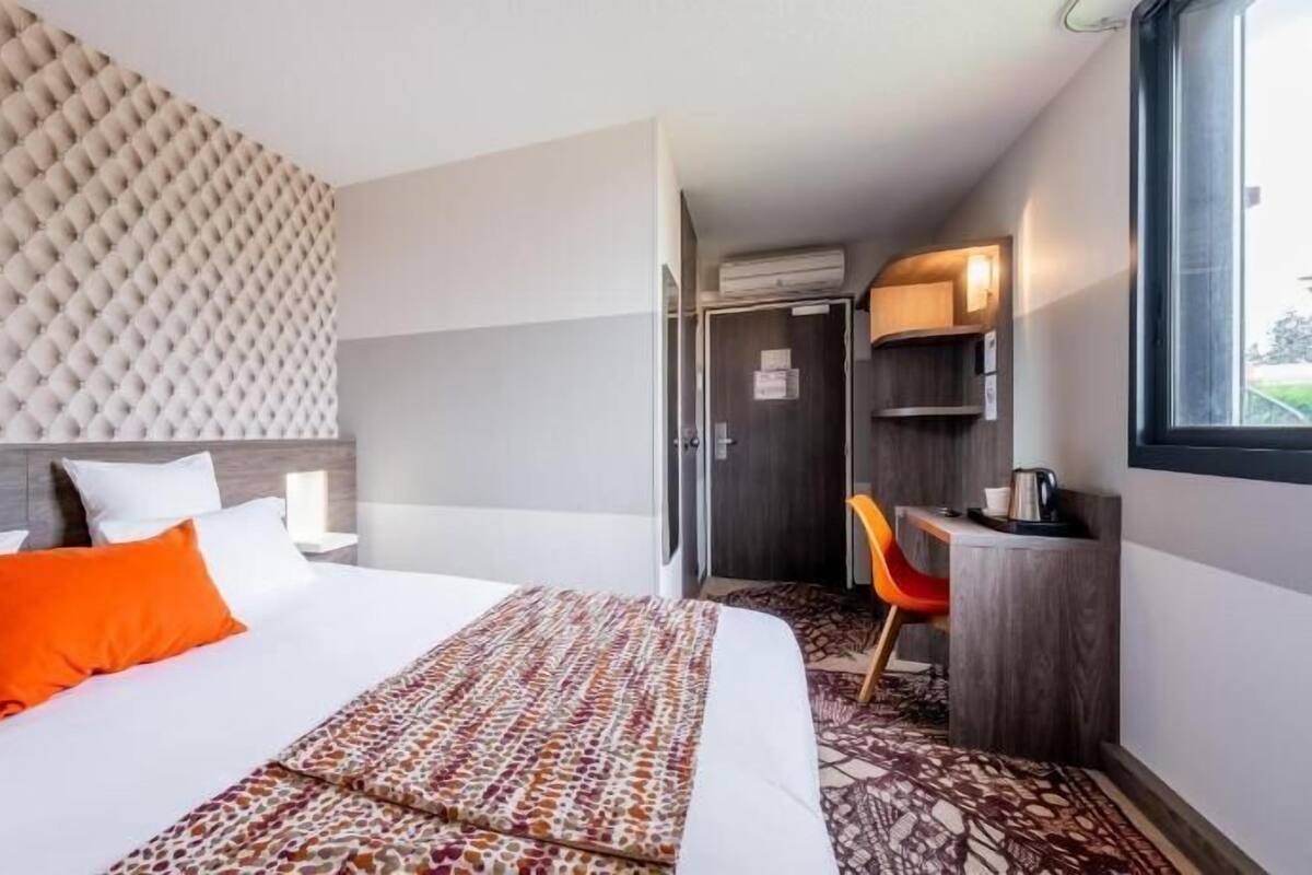 Ein Hotelzimmer mit einer gepolsterten Kopfteil, einem Bett mit weißer und orangefarbener Bettwäsche, einem gemusterten Überwurf, einem Schreibtisch mit Stuhl und einem Fenster.