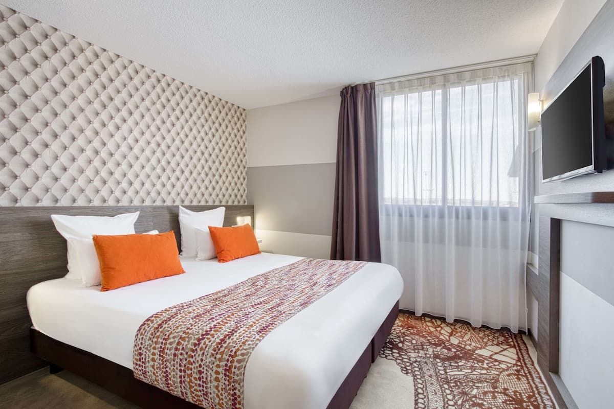 Ein Hotelzimmer mit einem großen Bett, einem an der Wand montierten Fernseher und einem Fenster mit Vorhängen.