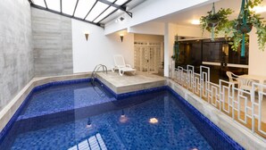 An indoor swimming pool with blue mosaic tiles, a white lounge chair, and a glass-enclosed area with hanging plants. - H Hotel (Balneário Camboriú)