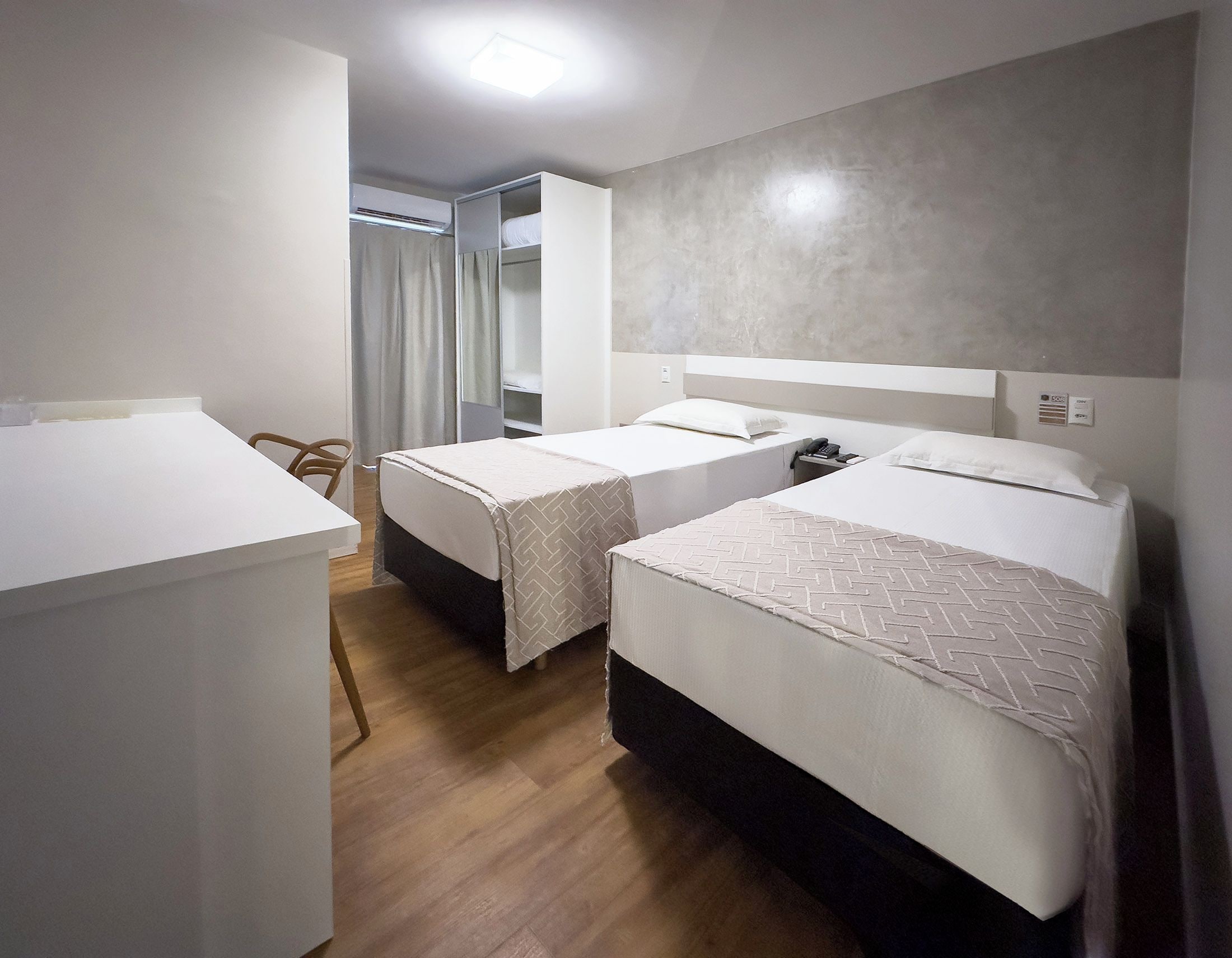 Quarto de hotel com duas camas de solteiro, uma escrivaninha e uma cadeira.