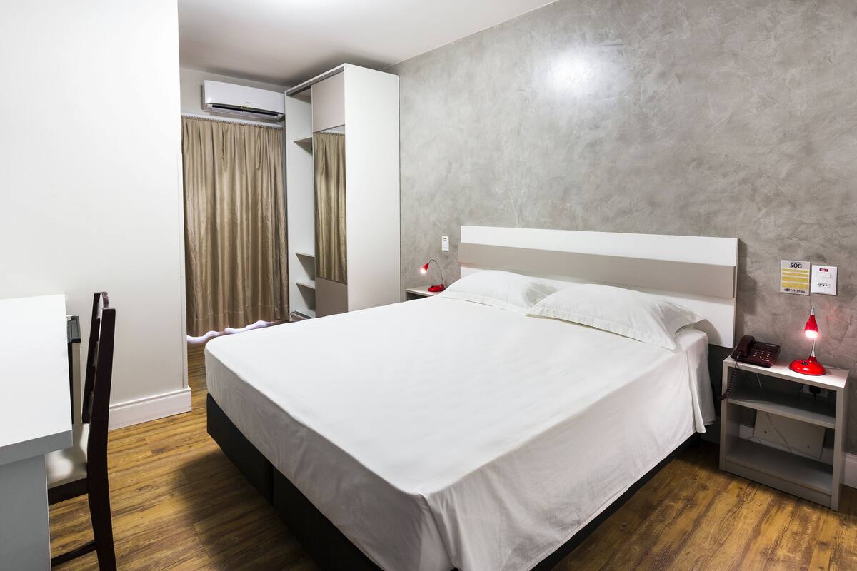 Una habitación de hotel moderna con una cama grande, una mesita de noche con lámpara, teléfono e interruptor de luz en la pared.