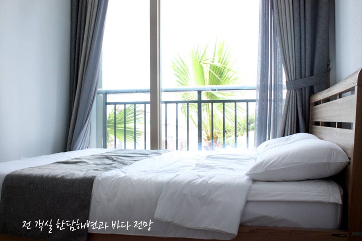 A bedroom with a bed, curtains, and a view of palm trees through a balcony railing.