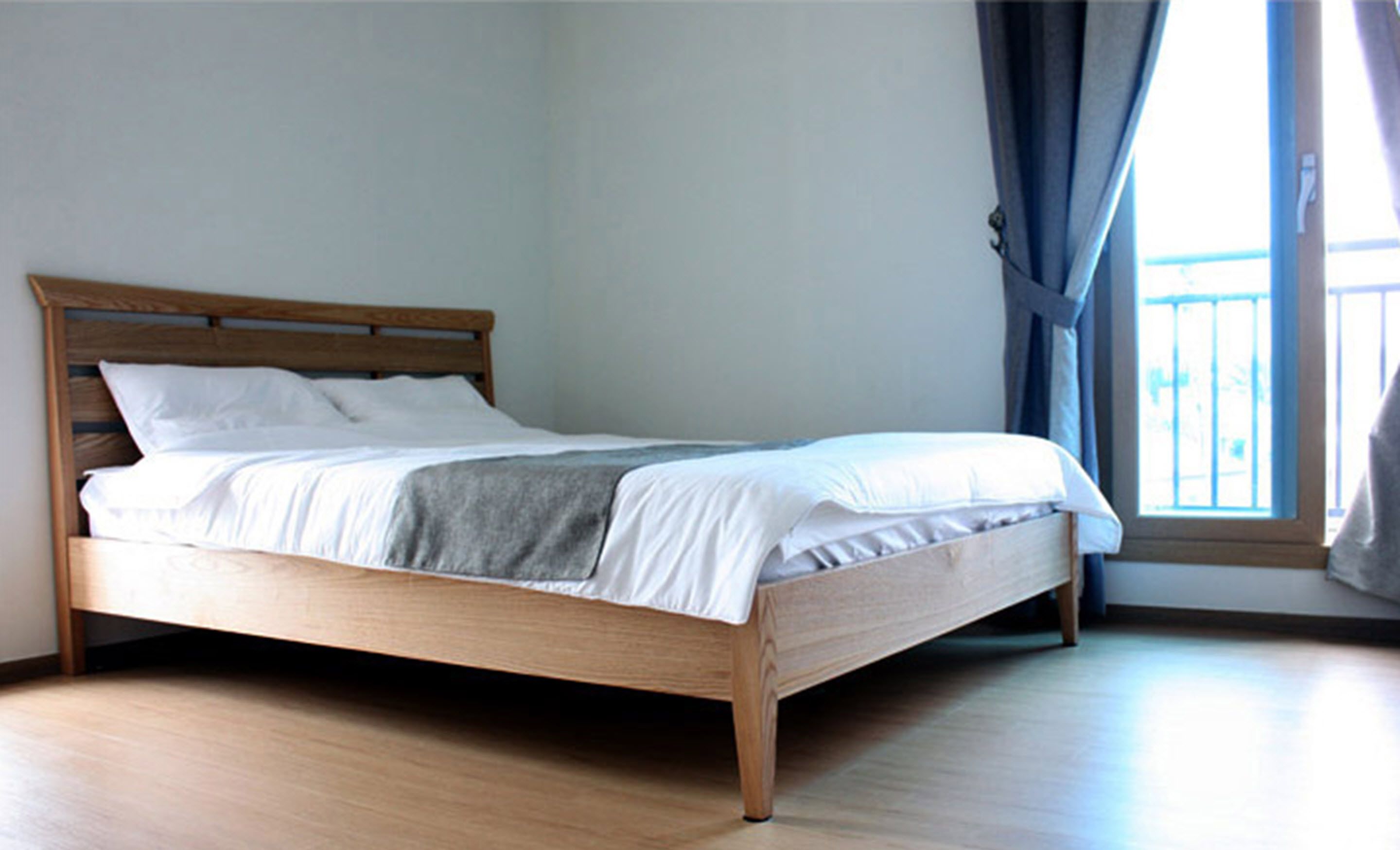 A single bed with a wooden frame and white bedding in a room with light-colored wooden flooring and a window with blue curtains.