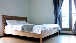 A single bed with a wooden frame and white bedding in a room with light-colored wooden flooring and a window with blue curtains. - Gilgaon Pension (Jeju City)