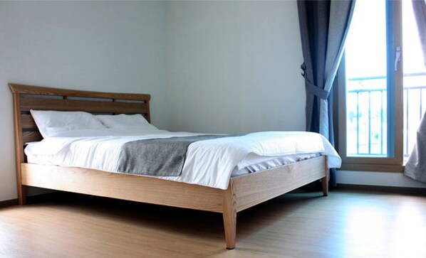 A single bed with a wooden frame and white bedding in a room with light-colored wooden flooring and a window with blue curtains. - Gilgaon Pension (Jeju City)