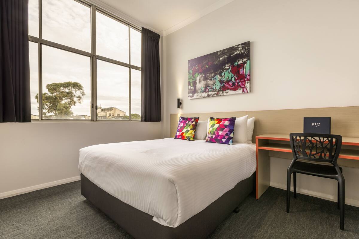 A hotel room with a large bed, a desk, and a chair. There is a window with curtains, a painting on the wall, and a view of trees and buildings outside.
