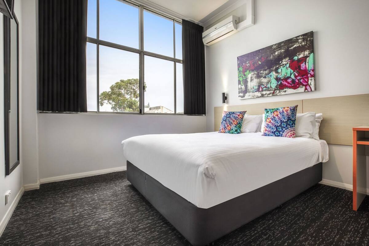 A hotel room with a large bed, a window with curtains, an air conditioning unit, and a painting on the wall.