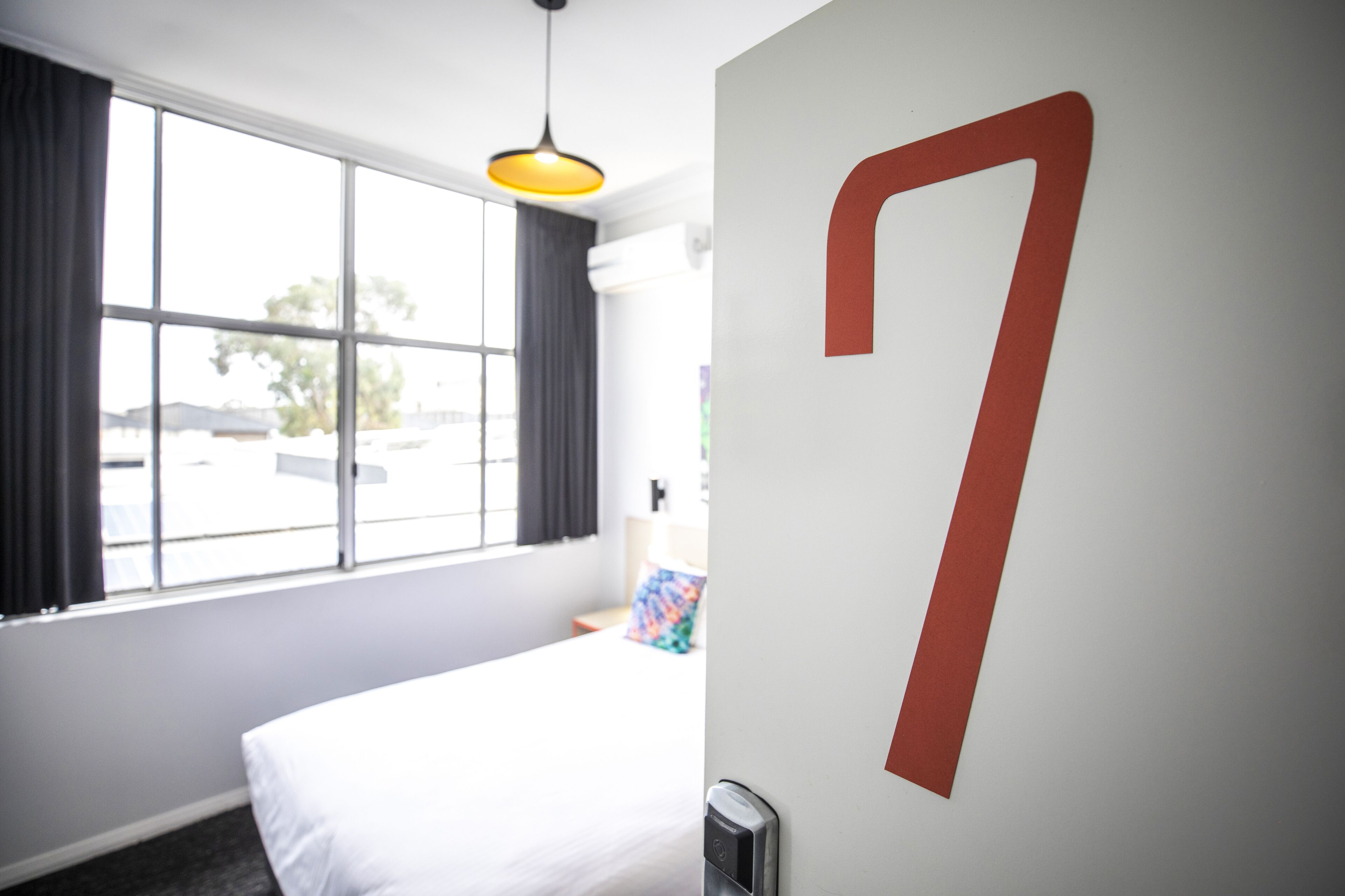 Une chambre d’hôtel avec une grande fenêtre, un lit, un numéro « 7 » fixé au mur et un luminaire suspendu.