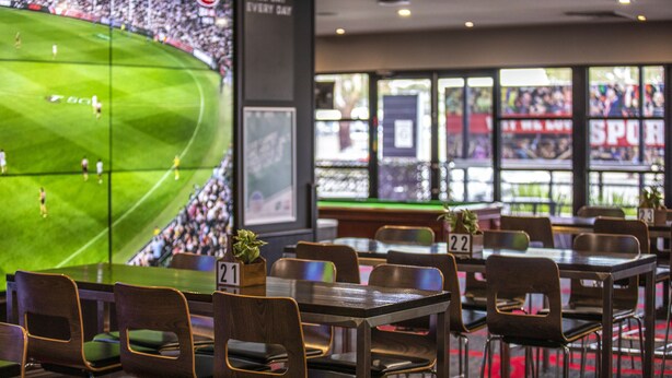 Un bar sportif doté de plusieurs écrans diffusant un match de football, de tables numérotées pour faciliter l’attribution des places, et d’une grande fenêtre offrant une vue sur le terrain.