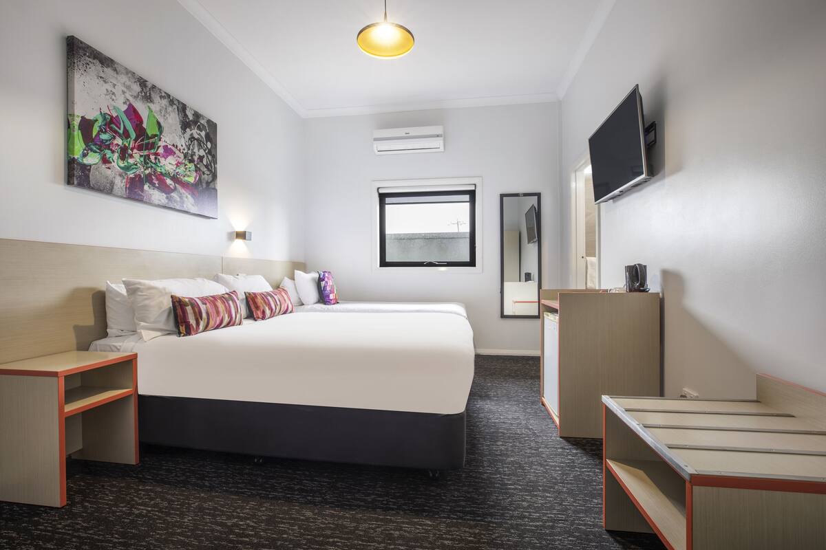 A hotel room with a large bed, bedside tables, a desk, a flat-screen TV, a window, and an air conditioning unit.