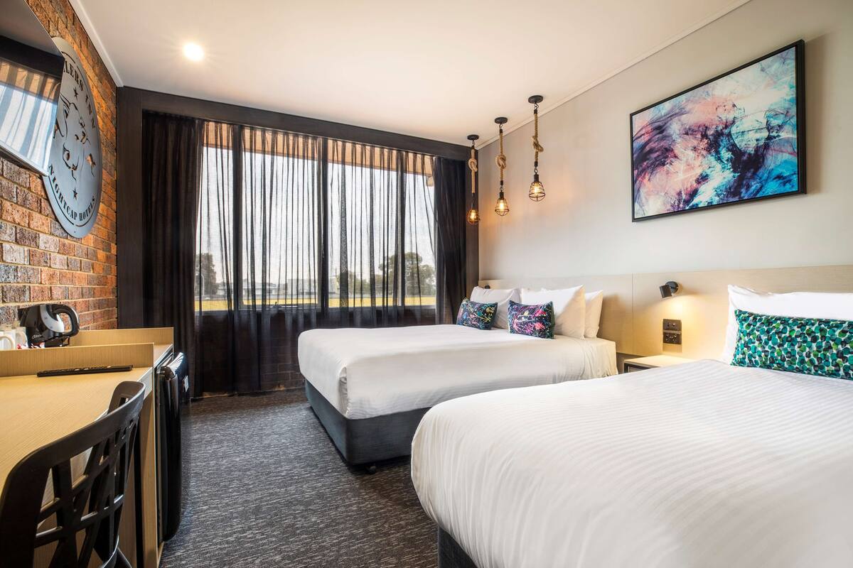 Ein Hotelzimmer mit zwei Betten, einem Schreibtisch, einem Stuhl, einer Uhr und einem großen abstrakten Gemälde an der Wand.