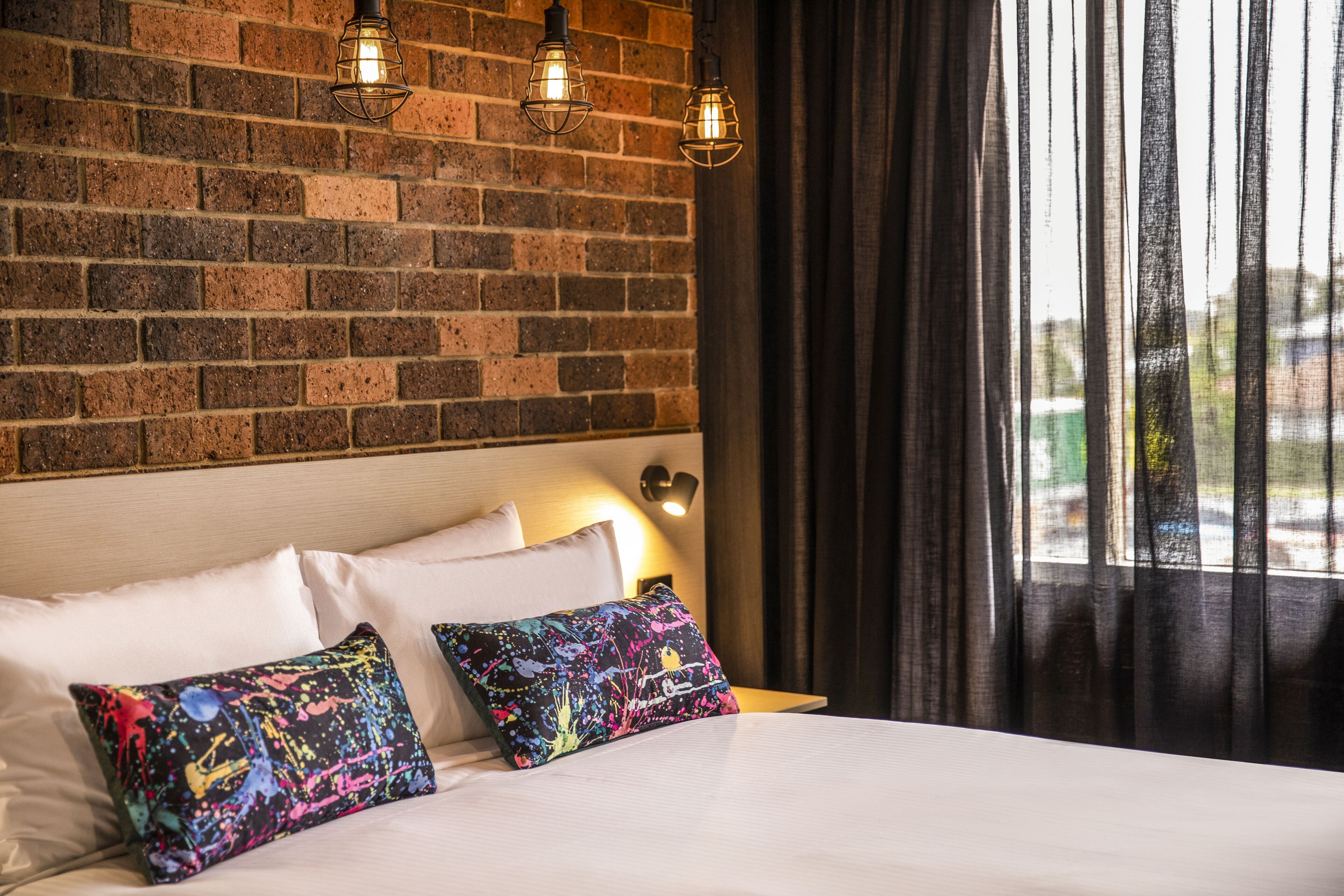 Ein Hotelzimmer mit einer Ziegelwand, einem Bett mit weißen Leinentextilien, bunten Kissen und einem Fenster mit Vorhängen.