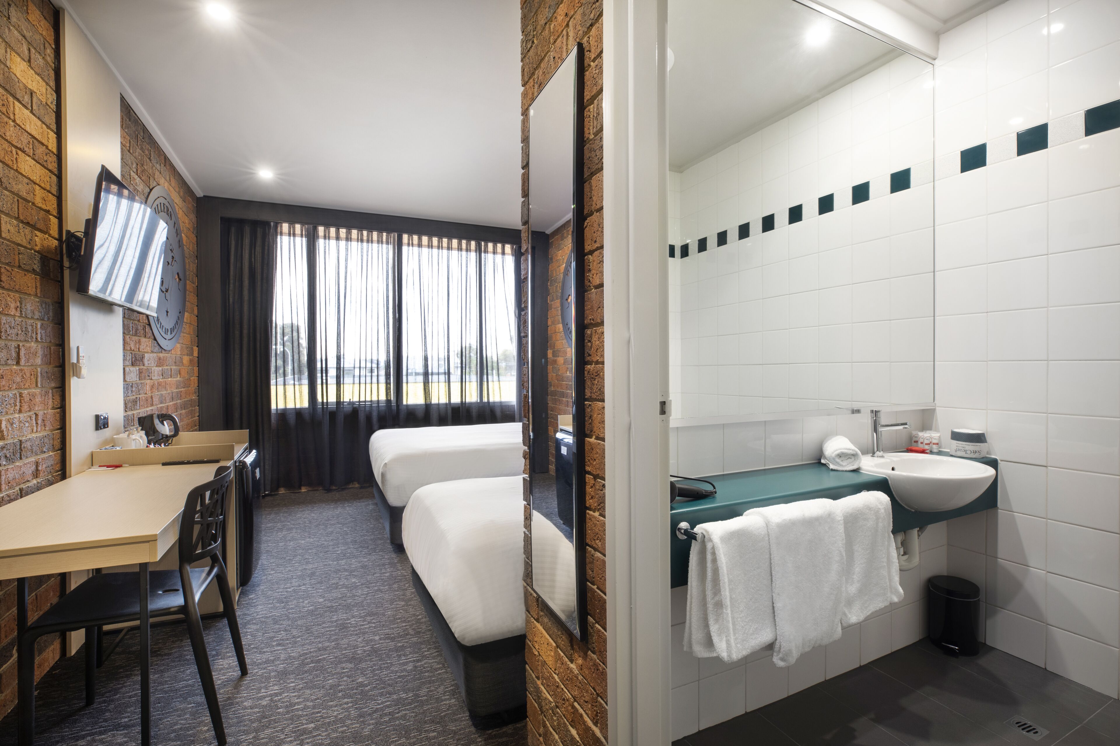 Ein Hotelzimmer mit Ziegelwand, Flachbildfernseher, Schreibtisch mit Stuhl, zwei Einzelbetten, Badezimmer mit Waschbecken und Spiegel sowie Blick aus dem Fenster.