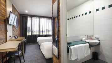 Ein Hotelzimmer mit Ziegelwand, Flachbildfernseher, Schreibtisch mit Stuhl, zwei Einzelbetten, Badezimmer mit Waschbecken und Spiegel sowie Blick aus dem Fenster.