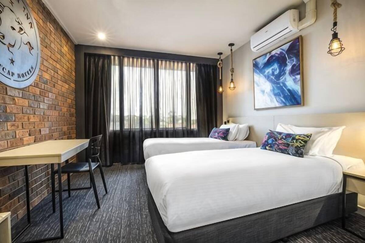 Ein Hotelzimmer mit zwei Betten, einem Schreibtisch, einem Stuhl, einer Wanduhr und einem großen Fenster mit Vorhängen.