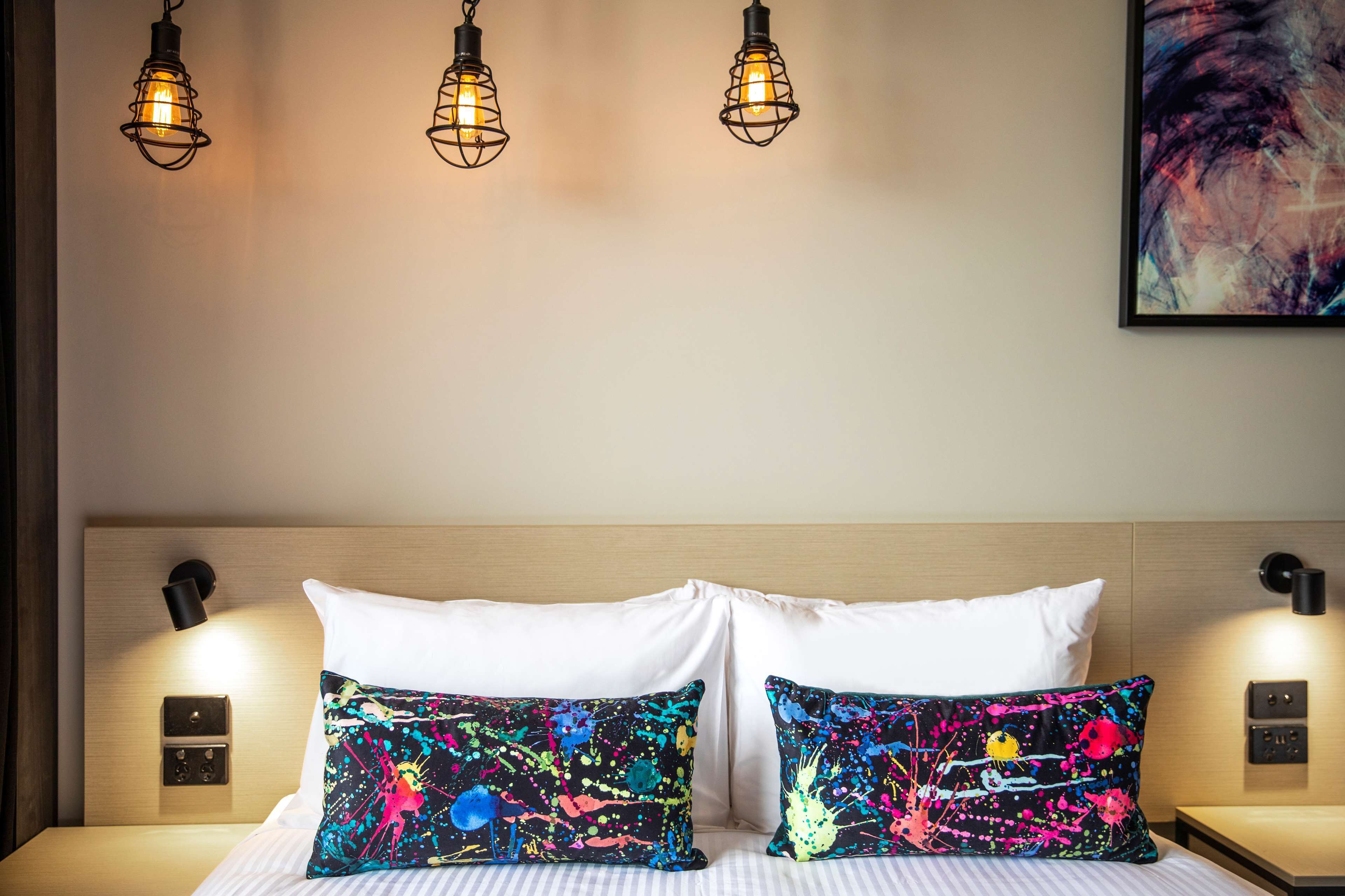 Ein Hotelzimmer mit Bett, zwei Kissen, Kopfteil, Nachttischlampen und einem Wandbild.